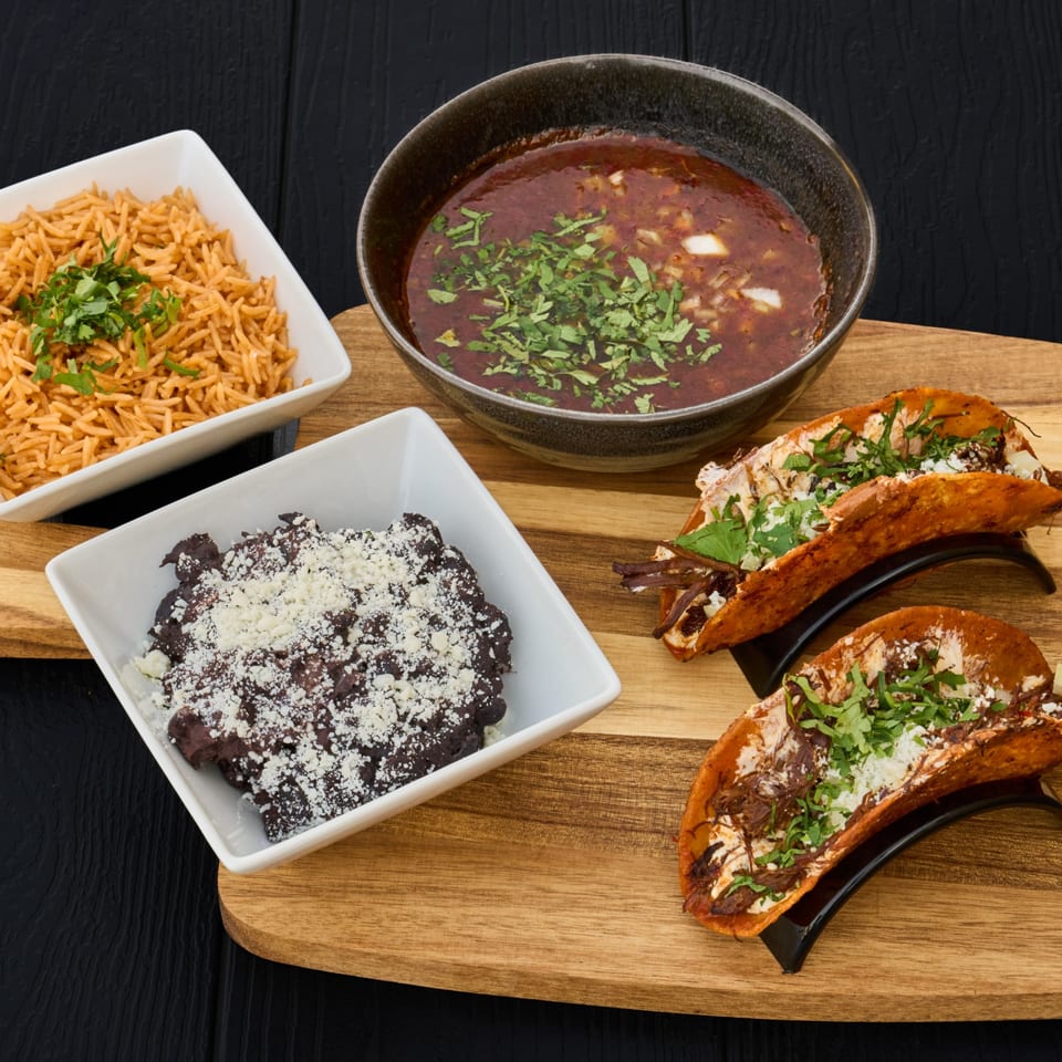Mexican meal featuring tacos, seasoned rice, black beans topped with cheese, and cilantro-garnished soup on a wooden serving board.