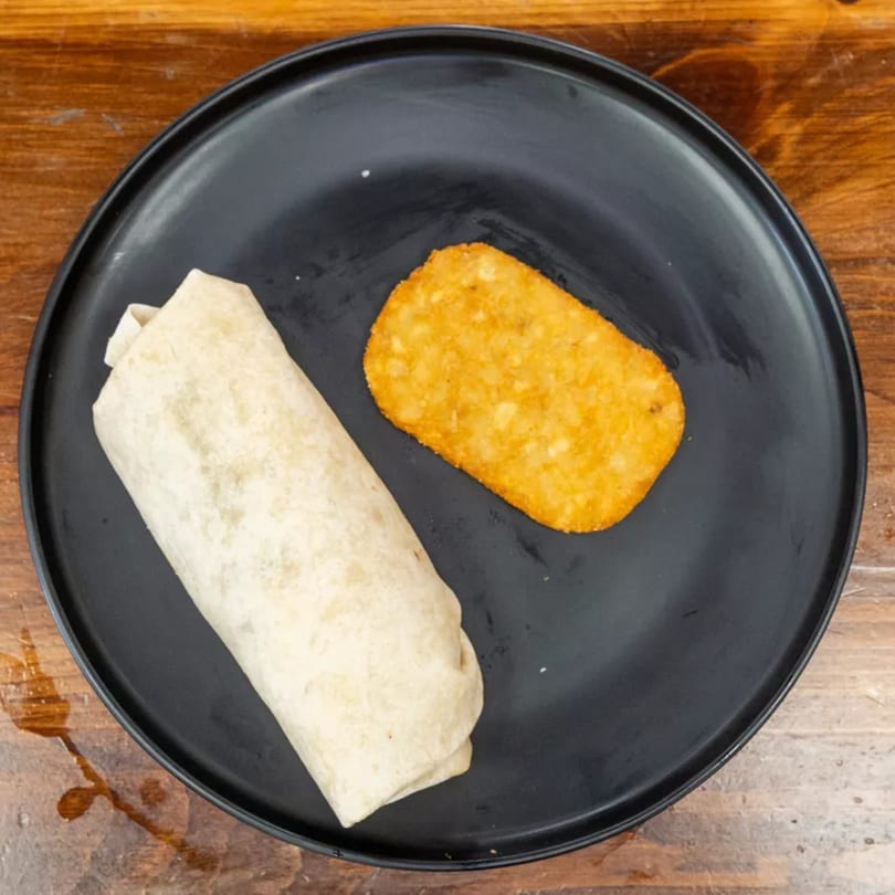 A breakfast burrito and a hash brown on a black plate, on a wooden table.