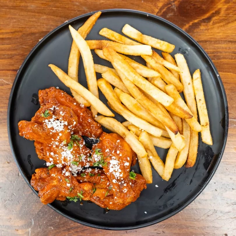 Spicy chicken wings with white cheese and parsley, served with golden french fries on a black plate.