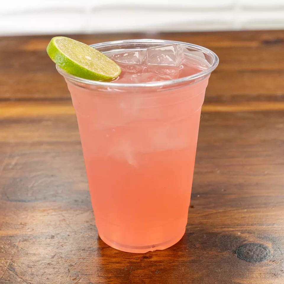 Pink lemonade with ice and a lime wedge in a clear plastic cup on a wooden table.