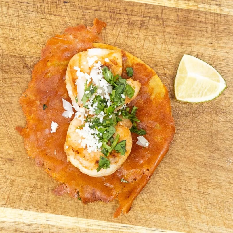 Shrimp taco with cheese, cilantro, and onions on a wooden board with a lime wedge.