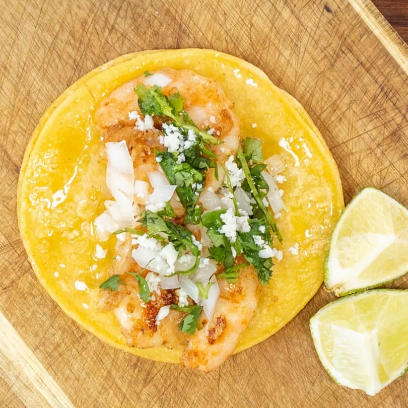 Overhead view of a shrimp taco with cilantro, onions, and cheese, with lime wedges on a wooden board.