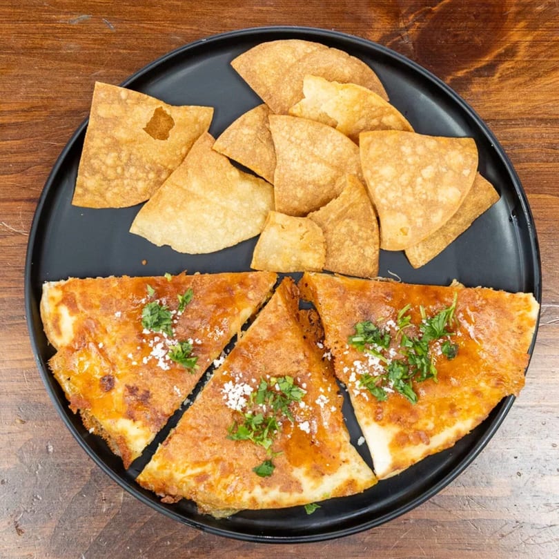 A black plate holds three slices of quesadilla topped with cilantro and powdered sugar, alongside tortilla chips.