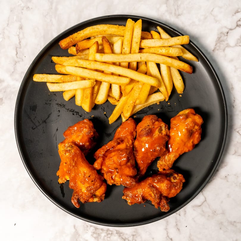 A black plate holds crispy french fries and several buffalo chicken wings on a marble surface.
