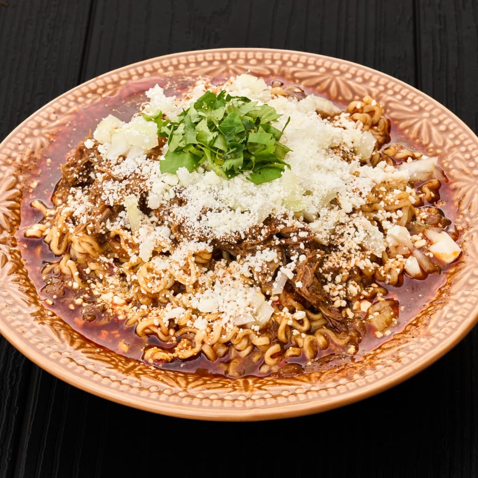 Pasta topped with shredded meat, crumbled white cheese, chopped herbs, onions, and nuts on an ornate plate.