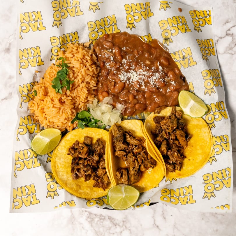 Three carne asada tacos with rice, beans, and lime wedges on Taco King branded paper.