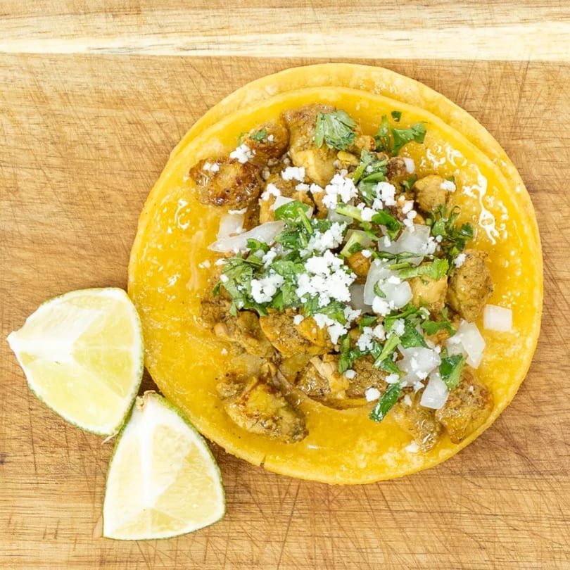 Chicken taco with cilantro, onions, and cotija cheese, served with two lime wedges on a wooden board.