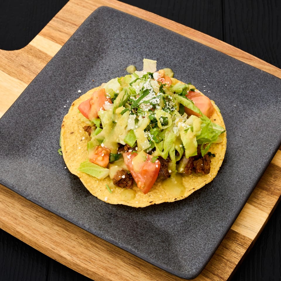 Crispy tostada topped with seasoned meat, lettuce, tomatoes, avocado, cilantro, creamy sauce, and crumbled cheese on a black plate.
