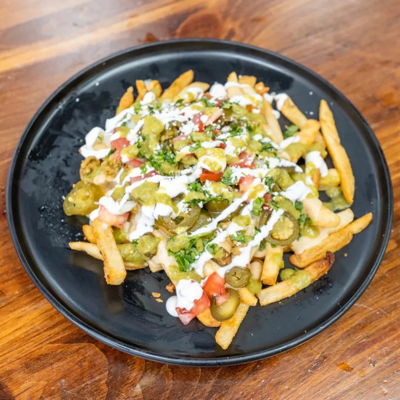 Loaded fries topped with green sauce, white sauce, jalapeños, tomatoes, and cilantro on a black plate.