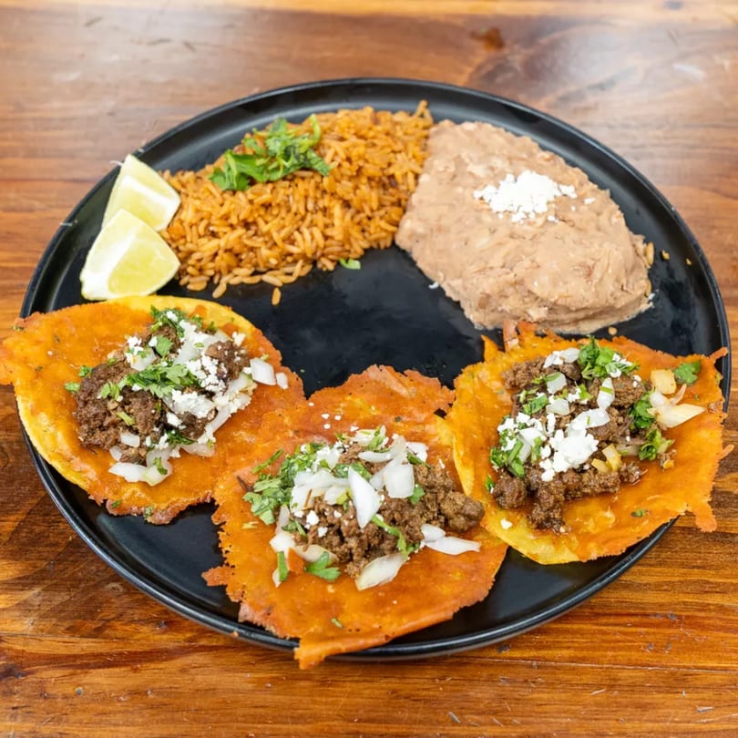 Three beef tacos with rice, refried beans, and lime wedges on a black plate.