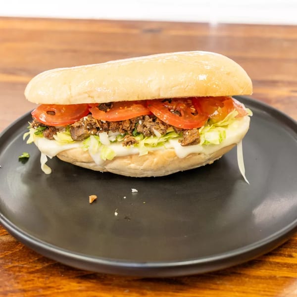 A close-up of a Mexican torta sandwich with meat, tomatoes, lettuce, and cheese on a black plate.