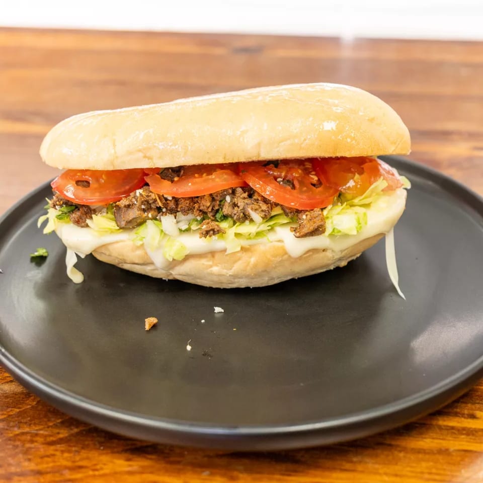 A close-up of a Mexican torta sandwich with meat, tomatoes, lettuce, and cheese on a black plate.
